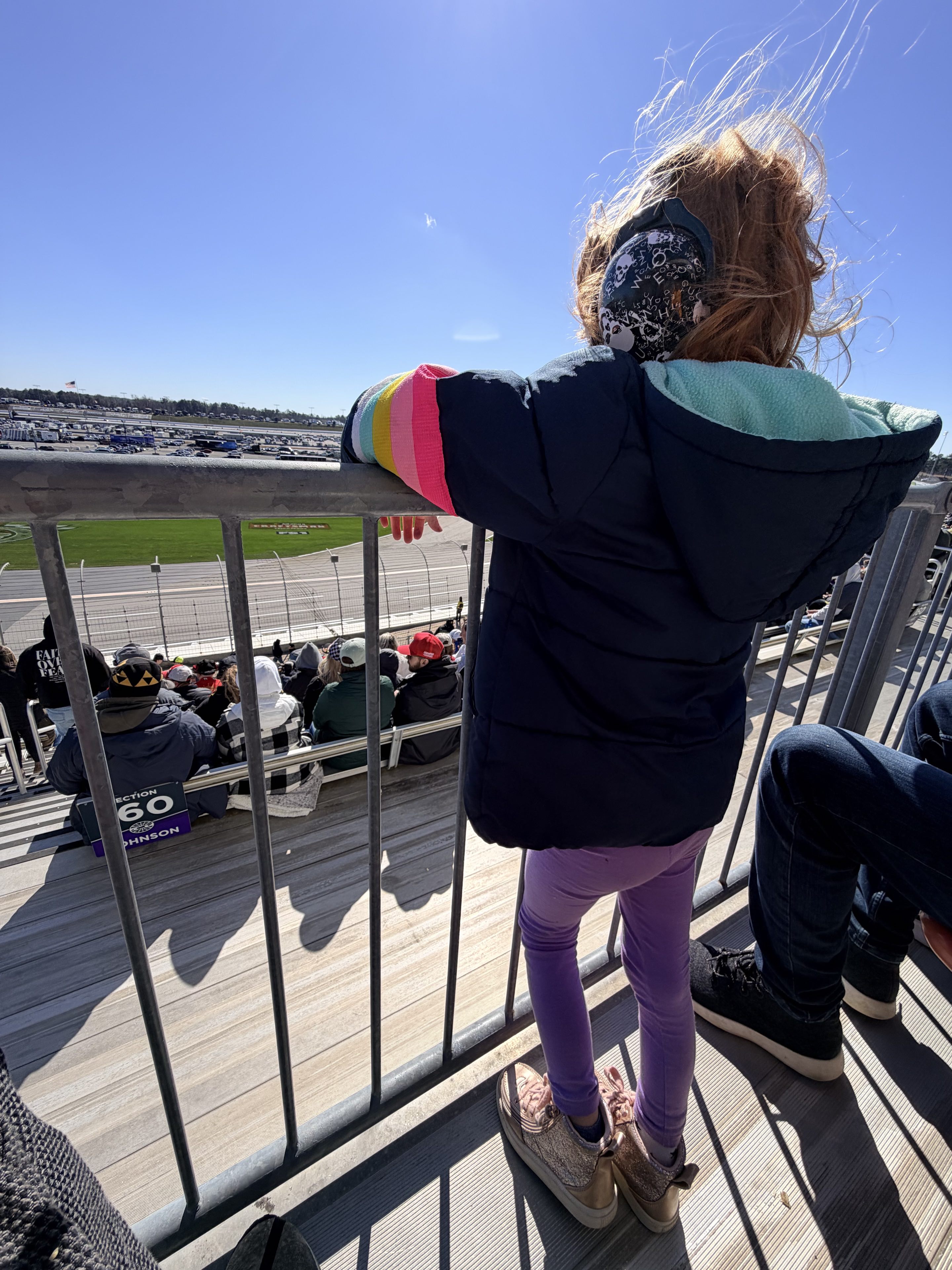 A kid with messy red hair, purple pants, a puffy jacket, and skull-and-crossbones over-ear hearing protection standing at a guardrail in the grandstands overlooking a race track